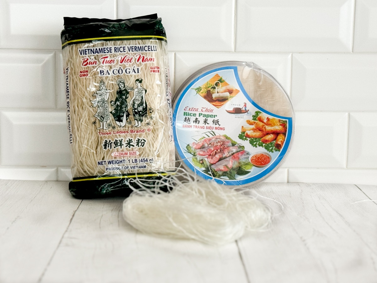 A package of dried vermicelli rice noodles and a package of round rice paper sheets on top of a white plank board with a white brick background.
