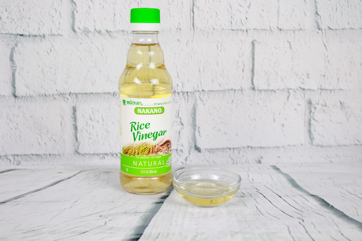 A bottle of Nakano rice vinegar and a small glass bowl with rice vinegar on a white plank board with a white brick background.