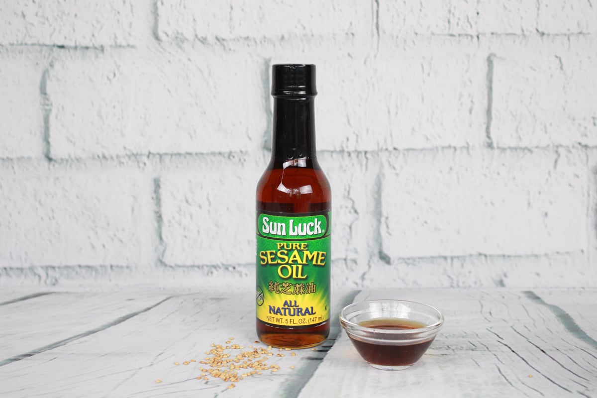 A bottle of Sun Luck sesame oil and a small glass bowl filled with sesame oil placed on top of a white plank board with white brick in the background.