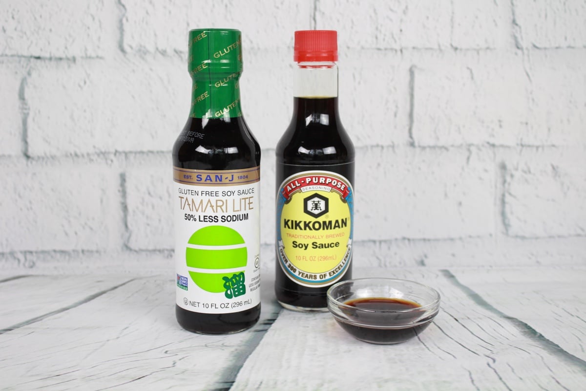 10 Essentials for Asian Cooking Two bottles of soy sauce and a small glass bowl filled with soy sauce placed on top of a white plank board with white brick in the background.