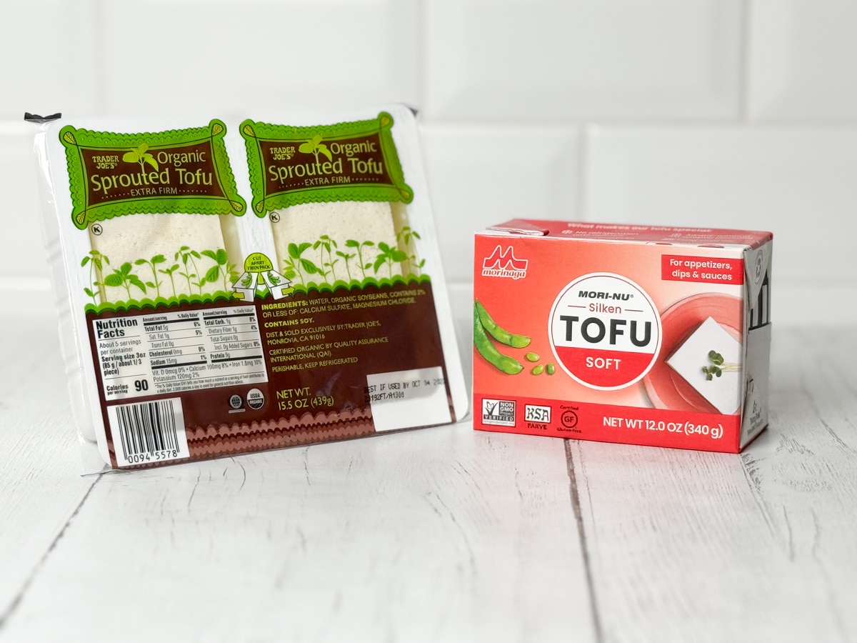 Two packages of tofu on top of a white plank board with a white brick background.
