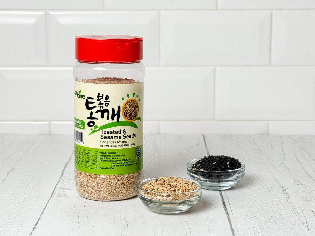 A plastic jar of toasted sesame seeds and two small bowls of white and black sesame seeds on the side on top of a white plank board with a white brick background.