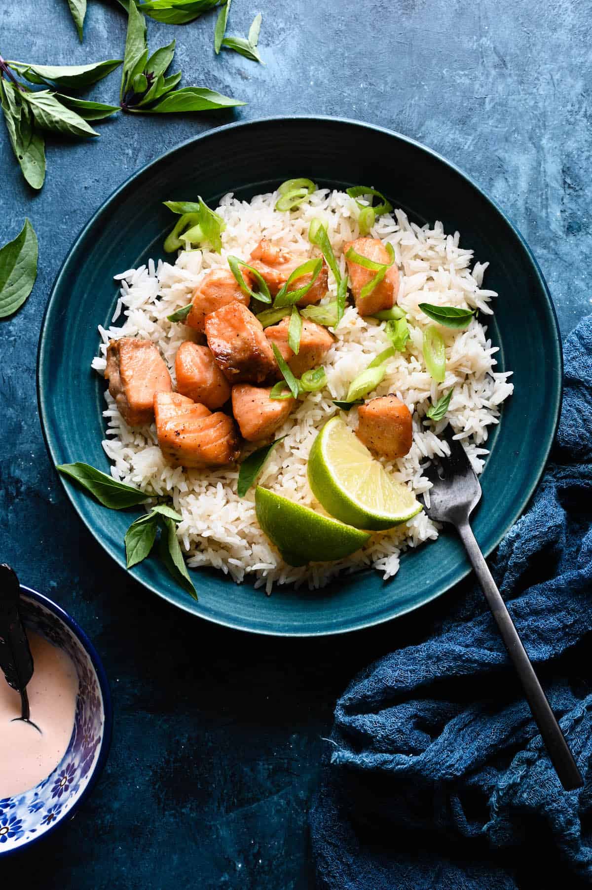 A healthy salmon recipe featuring crispy air fryer salmon bites on top of white rice with herbs and lime wedges in a round blue bowl with a fork on top of a blue surface.