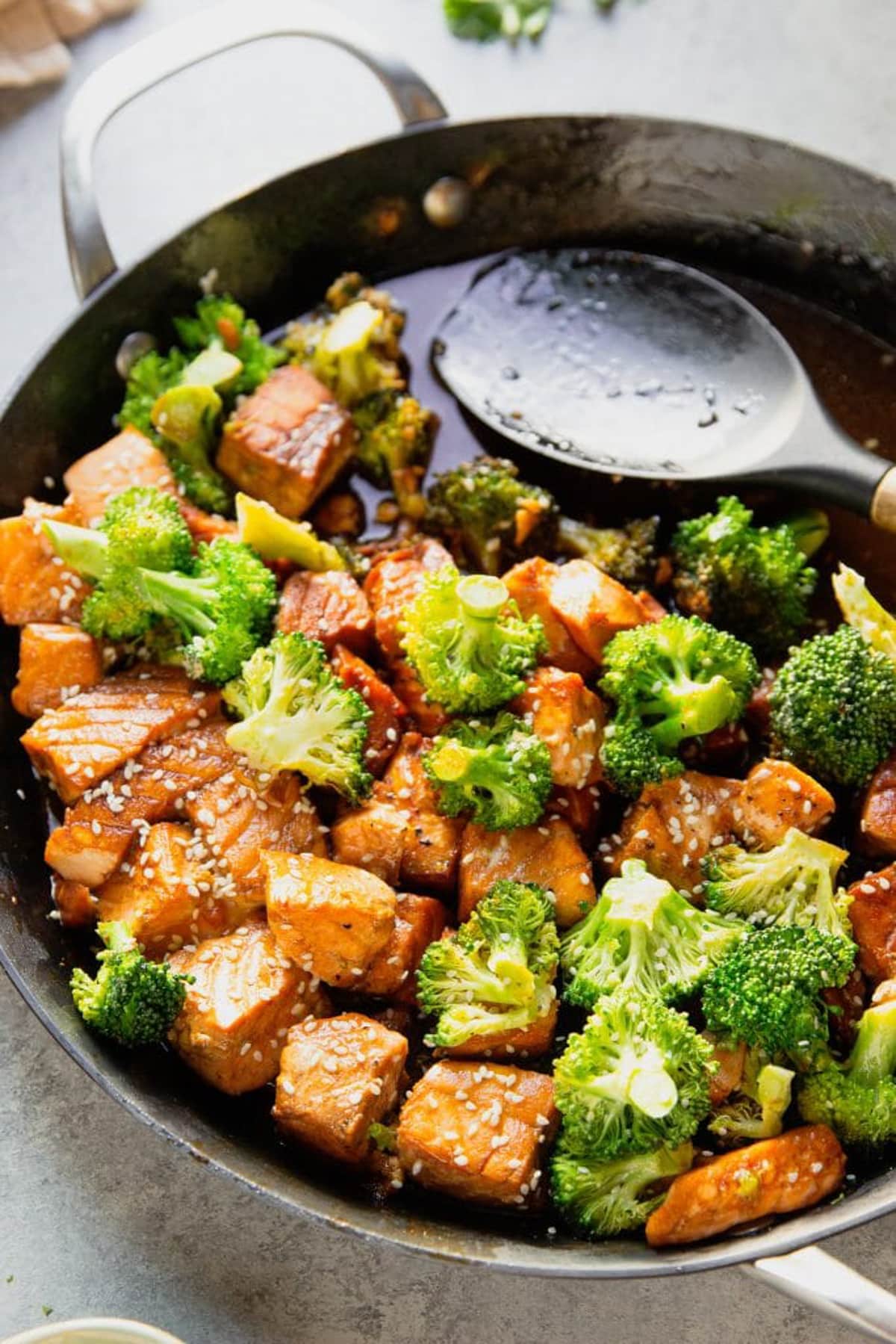 A healthy salmon recipe featuring a frying pan filled with a salmon and broccoli stir fry and a large serving spoon.