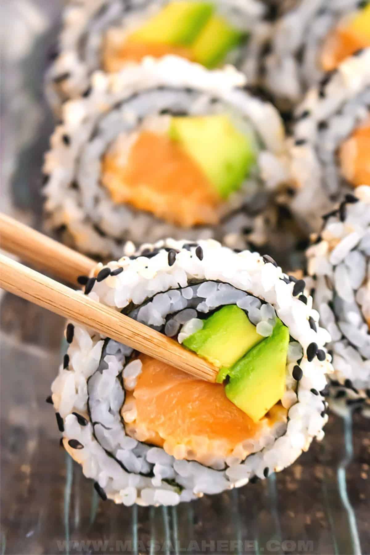 A healthy salmon recipe featuring a close-up image of a spicy salmon sushi roll being held up by a pair of chopsticks above the other pieces of sushi roll.