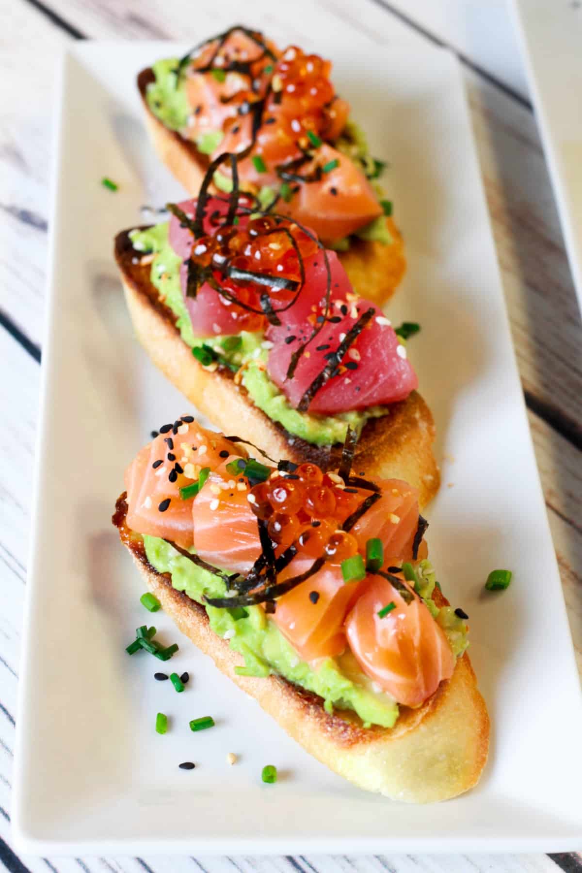 Sushi avocado toasts, a healthy salmon recipe featuring cubed sushi-grade salmon and tuna on a long white platter on top of a white wooden board.