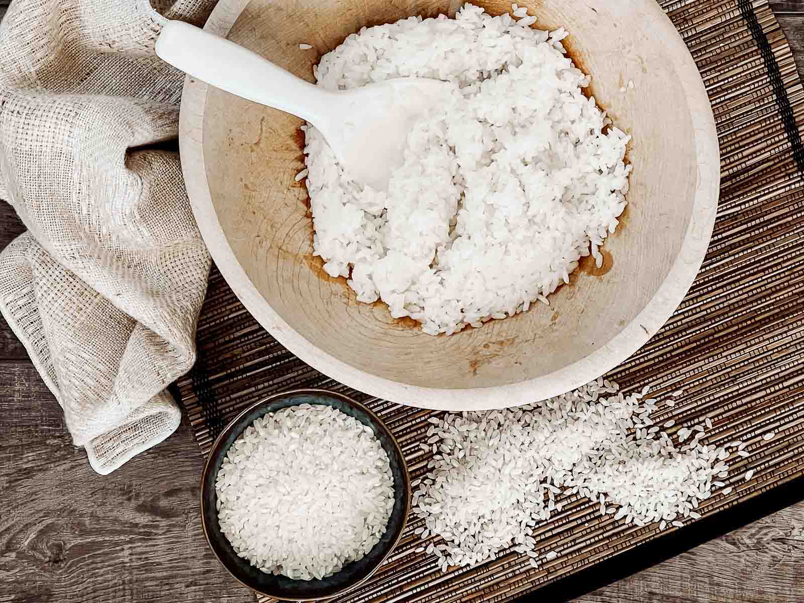 Cooked sushi rice in a large wooden with a white paddle inserted, and uncooked rice in a small bowl and on a bamboo mat on the side, on top of a wooden board with a linen napkin on the side.