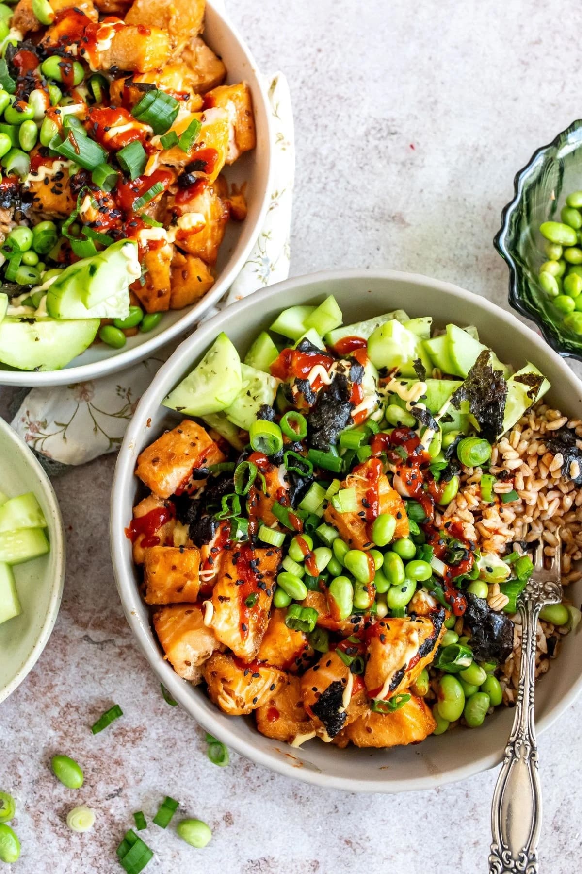 A healthy salmon recipe featuring two teriyaki salmon rice bowls on a marble surface with a bowl of edamame and cucumber on the side.