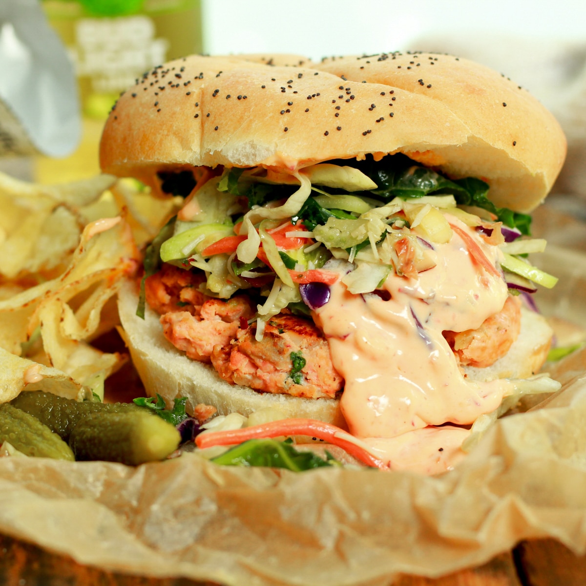 A healthy salmon recipe featuring a wild salmon burger topped with slaw and an aioli sauce on a piece of parchment paper with potato chips and pickles on the side.