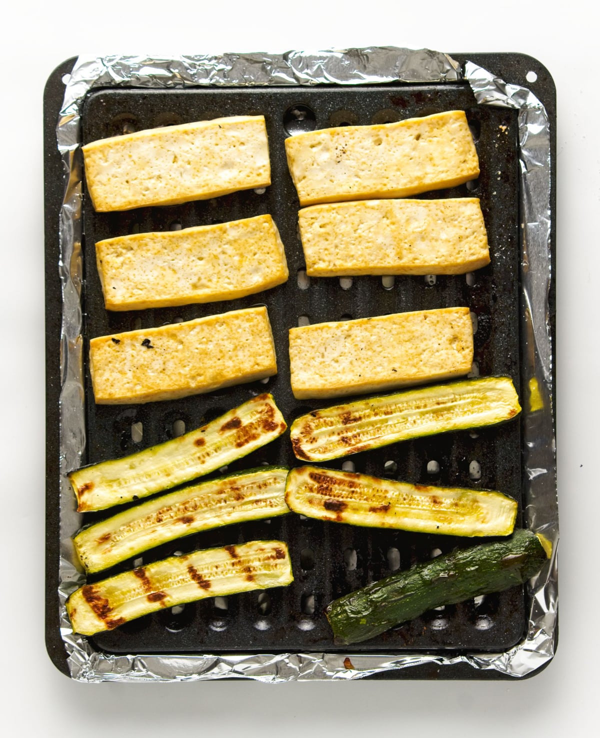 Baked tofu strips and zucchini on a black baking sheet lined with aluminum foil.