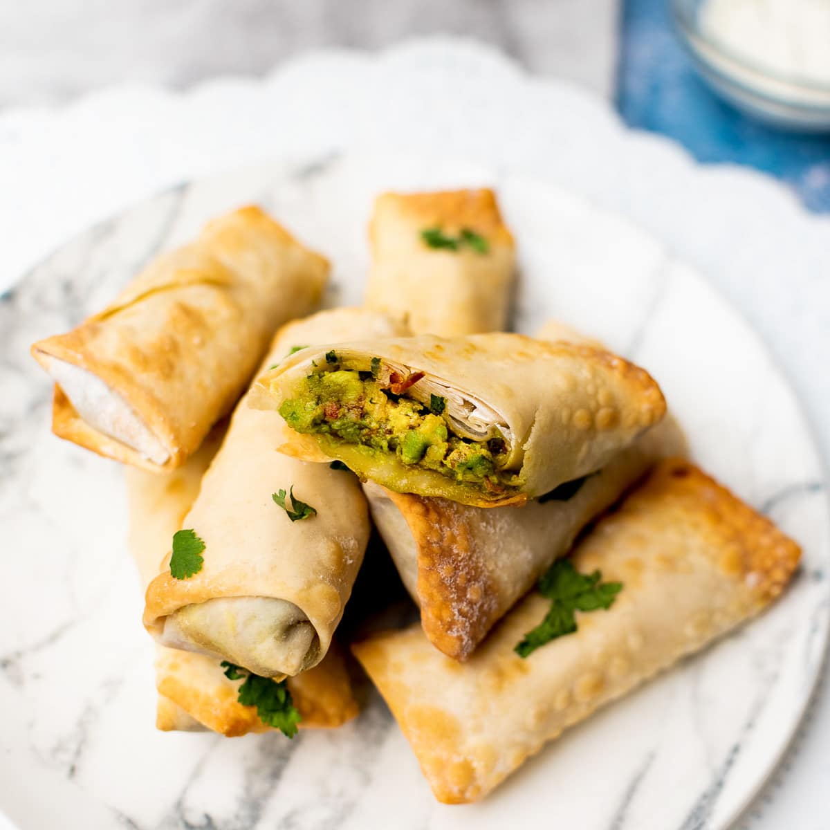 Air fryer Vegan Avocado Egg Rolls stacked on a white marbled plate on top of a blue board with a small bowl of sour cream in the background.