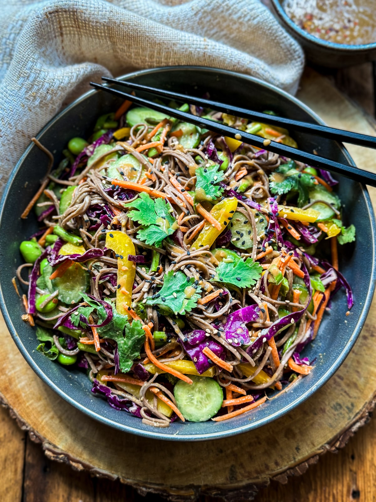A vibrant Vegan Asian Noodle Salad in a dark gray bowl topped with black chopsticks on a round wooden board with a linen napkin and small bowl of dressing on the side.